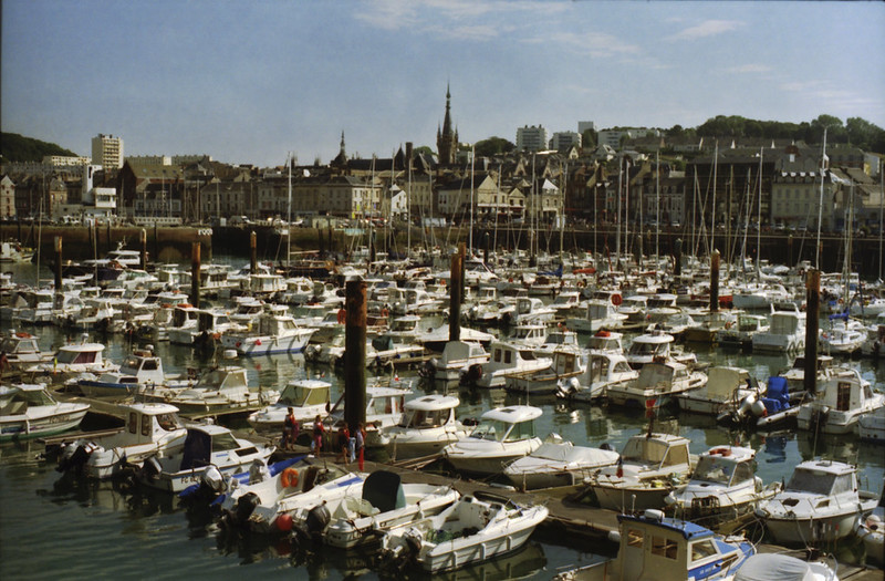 Matte gouache-style painting of a marina filled with many white boats and a town skyline with a single spire in the distance under a clear pale blue sky, calm daytime atmosphere.
