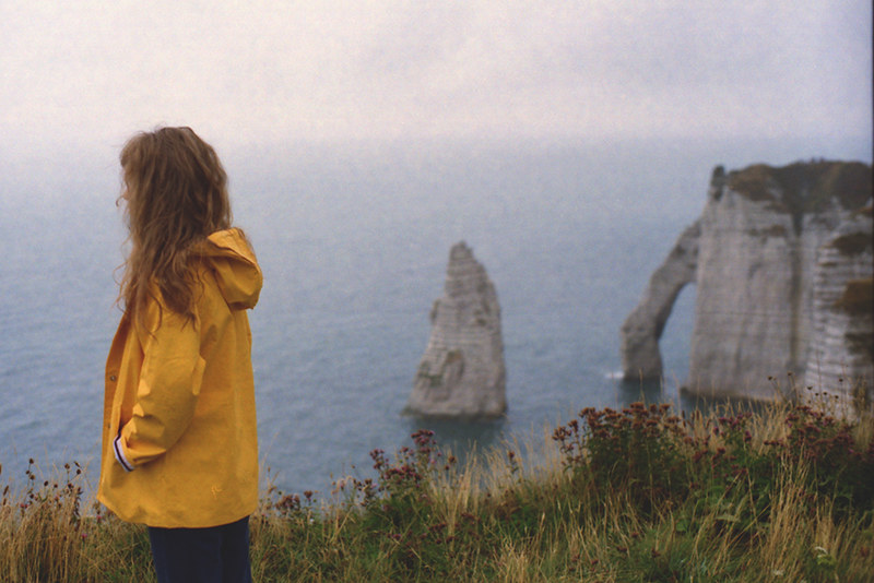 A solitary figure in a yellow raincoat looking out at sea cliffs and rock formations under an overcast blue-gray sky, painted in soft blues, grays, and a touch of mustard yellow, reflective mood, matte gouache poster style.